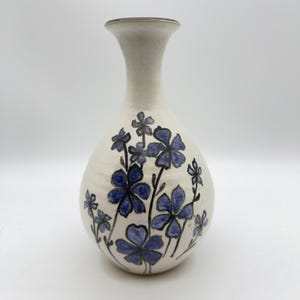 Könnte beinhalten: Eine Keramikvase mit cremefarbenem Grund und engem Hals. Der Körper der Vase ist mit handgemalten blauen Blumen mit dunklen Umrissen und Stielen verziert. Die Oberfläche hat eine subtile, strukturierte Optik mit einem dunklen Rand an der Öffnung.
