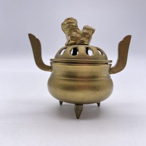 May include: A brass incense burner featuring a rounded body with three pointed feet and two upright handles. The removable lid has a series of arched cutouts and is topped with a sculpted figure of a mythical creature in a seated position.