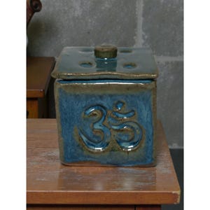 May include: A square ceramic container with a lid. The container is a mottled blue with a brown rim and a raised Om symbol on the front. The lid is a darker blue with a small knob on top.