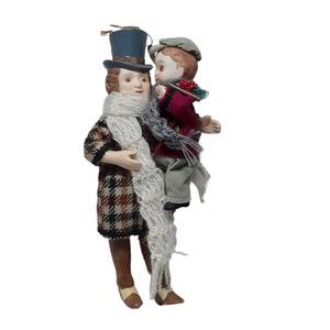 Puede incluir: Figura decorativa de una persona sosteniendo a un niño. La figura adulta lleva un sombrero de copa, un abrigo estampado y una bufanda blanca. El niño lleva un abrigo rojo y una bufanda gris. La figura es un adorno navideño.