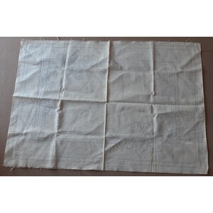 May include: A rectangular piece of off-white fabric with a floral design outlined in blue. The fabric is folded and has a raw edge on one side. The floral pattern is visible in the corners and center of the fabric.