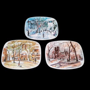 May include: Three ceramic plates with painted scenes of Parisian streets. The plates are rectangular and feature a white background with colorful illustrations of buildings, trees, and people. The top plate shows a street scene with a building in the background, the middle plate shows a street scene with a church in the background, and the bottom plate shows a street scene with a building in the background.