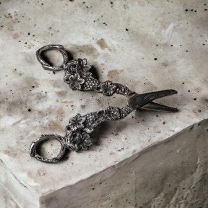 May include: Silver-toned decorative scissors with a floral design. The scissors have a vine-like pattern with leaves and flowers.
