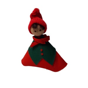 May include: A small, red felt doll with a green felt cape and a red knitted hat. The doll has a painted wooden face with brown hair and brown eyes.