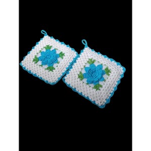 May include: Two square, crocheted potholders. Each potholder features a central blue flower with green leaves, surrounded by a white background and a blue border. A small blue loop is attached for hanging. The potholders are handmade.