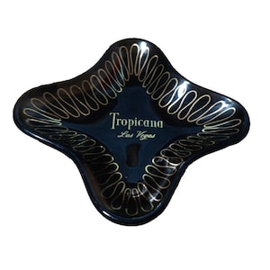 Op de afbeelding: Zwarte asbak met een klaverblad vorm en goudkleurige decoratieve accenten. De tekst "Tropicana Las Vegas" is in goud gedrukt. De asbak heeft een kleine inkeping om items op te plaatsen.