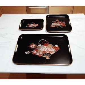 May include: A set of three black serving trays with gold trim and handles. Each tray features a decorative fan design with red, white, and gold floral accents. The trays are rectangular and square.