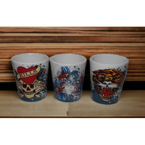 May include: Three white ceramic shot glasses with tattoo-style designs. One features a heart, skull, and the words "Kills Slow". Another has koi fish, and the third displays a tiger's head. Each has a blue splatter background.