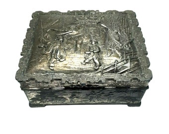 May include: A silver metal trinket box with an embossed design of people and an elephant. The box has a hinged lid and a detailed design around the edges.