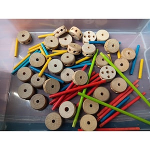 May include: A collection of wooden construction toy pieces including circular discs with center holes and various side perforations, alongside thin cylindrical rods in yellow, blue, red, and green, all contained within a clear plastic storage bin.