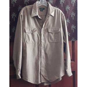 May include: A tan-colored long-sleeve button-down shirt with two chest pockets. The shirt has a collar and is hanging on a hanger. The brand name "J.Crew" is visible on the label.