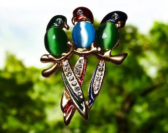May include: A gold-toned brooch featuring three parrot figures perched on a branch. The parrots have green, blue, and red accents, with gemstone-like bodies. The branch and tail feathers are embellished with clear crystals.