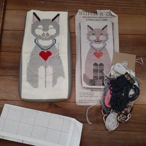 Può includere: Un kit di punto croce per un astuccio per occhiali che presenta un gatto grigio con un cuore rosso. Il kit include tessuto Aida preassemblato, filo da ricamo e una cerniera. Il kit è etichettato "Stitch & Zip Preassembled Cross Stitch Eyeglass Case 57603X Cat/Heart".