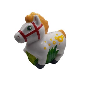 Może przedstawiać: Biała ceramiczna skarbonka w kształcie konia z czerwono-białym uzdą, żółtą grzywą i ogonem, zieloną trawą i żółtymi kropkami na ciele.