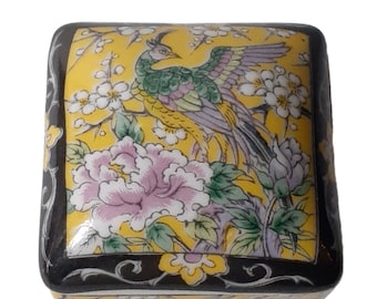 May include: A small, square, yellow ceramic trinket box with a black border. The box is decorated with a colorful floral design featuring a peacock with a long tail, pink flowers, and green leaves.