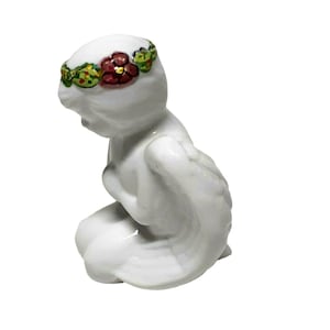 Puede incluir: Figurita de porcelana blanca de un ángel arrodillado con alas. El ángel tiene una corona de flores alrededor de la cabeza con flores rojas, verdes y amarillas.