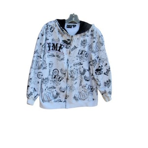Op de afbeelding: Witte hoodie met een zwarte rits en zwarte print over de hele voorkant. De print bevat de letters "FMF" en andere ontwerpen.