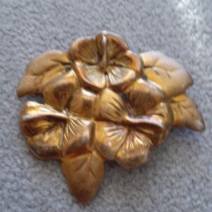 May include: A gold-colored metal floral brooch featuring a cluster of pansies and leaves. The brooch has a detailed, textured surface and a vintage aesthetic. The pansies are arranged in a layered design.