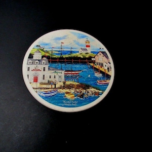 May include: A round ceramic coaster with a colorful painting of a seaside town. The scene includes a lighthouse, a dock, boats, and buildings. The text "Fenwick Harbor" is printed on the coaster.