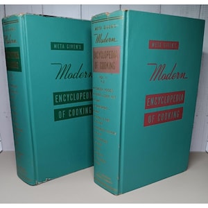 May include: Two vintage teal-colored cookbooks titled "Meta Given's Modern Encyclopedia of Cooking." The books have red and white text, with the title prominently displayed. The spines list volume numbers and cooking topics.