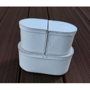 May include: Three white oval-shaped boxes with lids. The boxes are stacked, with the top box split into two sections. Each box has a contrasting stitch detail around the edges. The boxes are on a brown wooden surface.