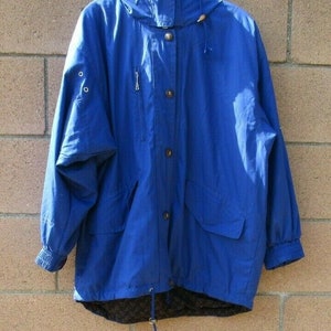 May include: A vibrant blue hooded jacket with a drawstring, zipper, and button closure. The jacket features two front pockets, elastic cuffs, and a dark lining. The jacket is hanging against a brick wall.