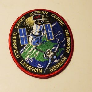 Puede incluir: Un parche bordado en blanco y negro con un borde rojo. El parche presenta un transbordador espacial y el telescopio espacial Hubble orbitando la Tierra. El texto en el parche dice "CAREY ALTMAN CURRIE GRUNSFELD LINNEHAN NEWMAN MASSIMINO".