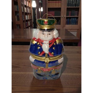 May include: A decorative ceramic nutcracker figurine in a soldier's uniform. The figurine is painted with blue, red, gold, and white details. It has a green hat and a gold-colored metal loop for hanging.