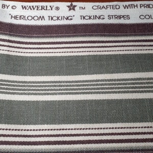 以下が含まれることがあります： 緑、白、茶色のストライプの生地のクローズアップ。生地には「'HERLOOM TICKING' TICKING STRIPES」という文字が印刷されています。