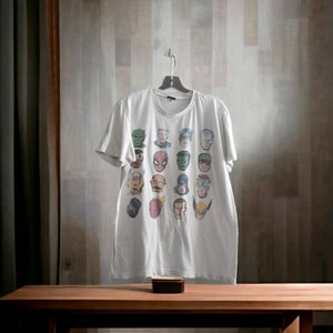 May include: White t-shirt featuring a colorful collage of superhero faces. The shirt is hanging on a hanger against a neutral background. A small, dark wooden box sits on a wooden table below the shirt.