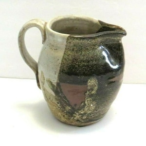 Puede incluir: Una jarra de cerámica marrón y blanca con un esmalte moteado. La jarra tiene un cuerpo redondeado y un asa.