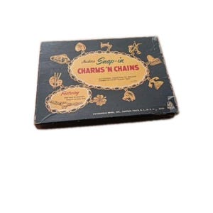 May include: Vintage Hasbro Snap-in Charms 'N Chains set. The box is dark blue with gold-colored text and illustrations of charms. The text reads "Featuring How easy to assemble Snap-in jewelry links."