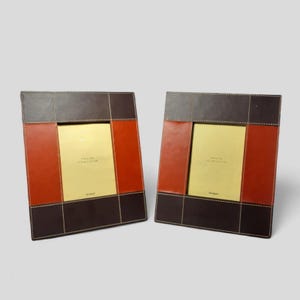 Puede incluir: Dos marcos de fotos de cuero marrón con detalles en cuero naranja. Cada marco tiene un borde cosido y un paspartú de color dorado. El texto "SMALL 177MM X 127MM" está impreso en el paspartú.