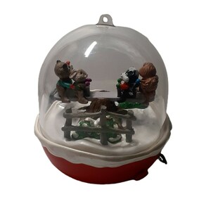 May include: A clear plastic snow globe ornament with a red base. Inside the globe, four small animal figurines are on a seesaw. The animals are a brown bear, a brown rabbit, a black and white dog, and a brown squirrel. The seesaw is made of wood and has a tree trunk as the base. There is a small wooden fence around the base of the seesaw.