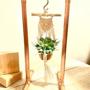 May include: A copper pipe plant stand with a macrame plant hanger and a small succulent plant. The stand has a hook on top and a wooden dowel across the top.