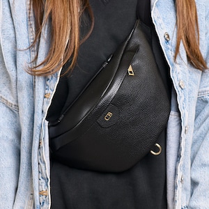 Peut inclure: Un sac banane en cuir noir grain&eacute; dot&eacute; d'une fermeture &eacute;clair dor&eacute;e et d'un anneau en D en m&eacute;tal dor&eacute; sur le c&ocirc;t&eacute;. L'avant du sac est emboss&eacute; avec les lettres dor&eacute;es CB. Le sac est port&eacute; en bandouli&egrave;re avec une sangle noire r&eacute;glable.