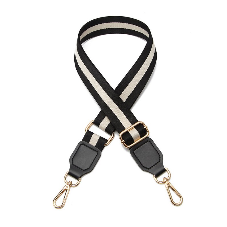 May include: A wide, adjustable bag strap featuring a black and beige striped pattern. The strap includes black faux leather end tabs and polished gold-tone metal hardware, including a sliding buckle and two swivel lobster claw clasps at the ends.