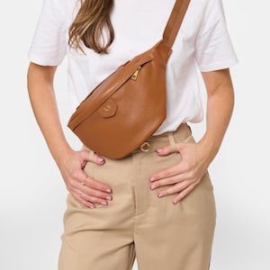 Peut inclure: Un sac banane en cuir grain&eacute; marron clair dot&eacute; d'une fermeture &eacute;clair dor&eacute;e, d'une sangle en tissu marron r&eacute;glable et d'un petit &eacute;cusson gaufr&eacute; sur le devant avec les initiales KS. Le sac est port&eacute; en bandouli&egrave;re sur un t-shirt blanc et un pantalon beige.
