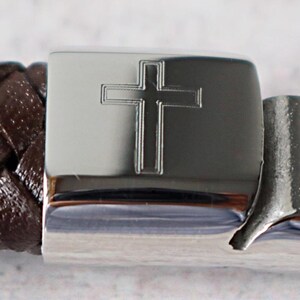 May include: A brown leather bracelet with a silver clasp featuring an engraved cross.