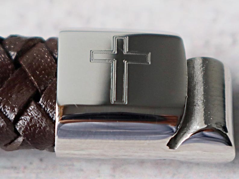 May include: A brown leather bracelet with a silver clasp featuring an engraved cross.