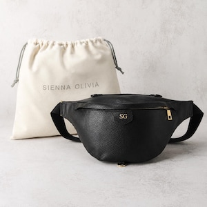 Peut inclure: Un sac banane noir &agrave; texture grain&eacute;e avec une fermeture &eacute;clair dor&eacute;e et les initiales SG dor&eacute;es en relief sur le devant. Derri&egrave;re le sac se trouve un pochon en tissu cr&egrave;me avec cordon de serrage et le texte SIENNA OLIVIA imprim&eacute; en gris.