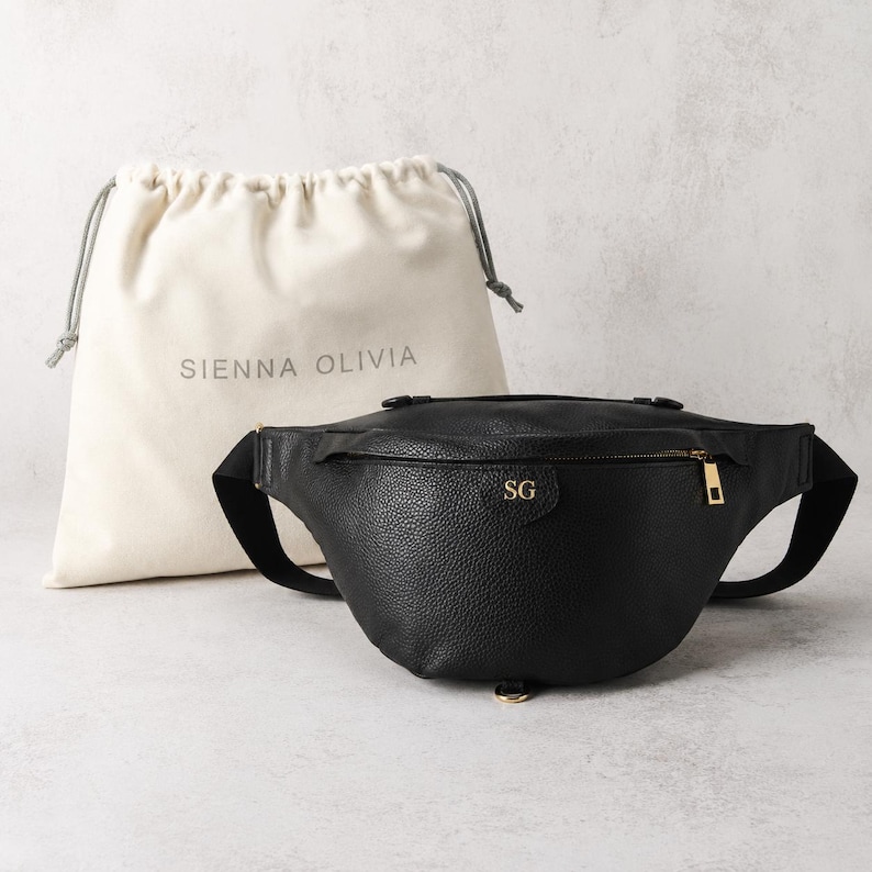 Peut inclure: Un sac banane noir &agrave; texture grain&eacute;e avec une fermeture &eacute;clair dor&eacute;e et les initiales SG dor&eacute;es en relief sur le devant. Derri&egrave;re le sac se trouve un pochon en tissu cr&egrave;me avec cordon de serrage et le texte SIENNA OLIVIA imprim&eacute; en gris.