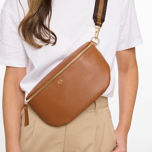 May include: A tan pebbled texture crossbody bag with a gold zipper closure and the gold embossed initials KS on the front. The bag features a wide, adjustable brown and tan striped fabric strap attached with gold metal hardware clips.