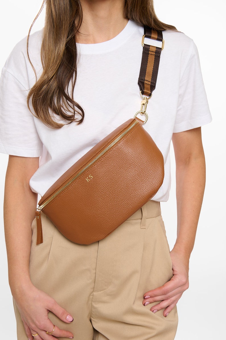 May include: A tan pebbled texture crossbody bag with a gold zipper closure and the gold embossed initials KS on the front. The bag features a wide, adjustable brown and tan striped fabric strap attached with gold metal hardware clips.
