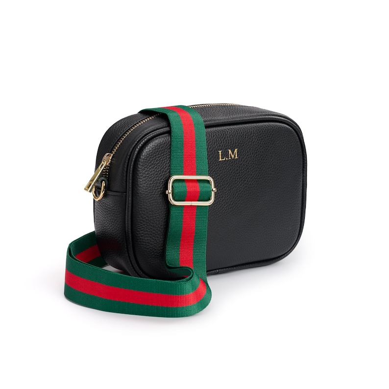 May include: A black pebbled leather camera bag featuring gold-tone hardware, a top zipper closure, and the gold-embossed initials L.M. on the front. The bag includes an adjustable, detachable woven strap with a green and red striped pattern.