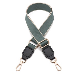 May include: A sage green adjustable bag strap featuring a woven texture with light beige edges. The strap includes black faux leather tabs at both ends, connected to gold-toned metal hardware, including a sliding buckle and two swivel lobster clasps.