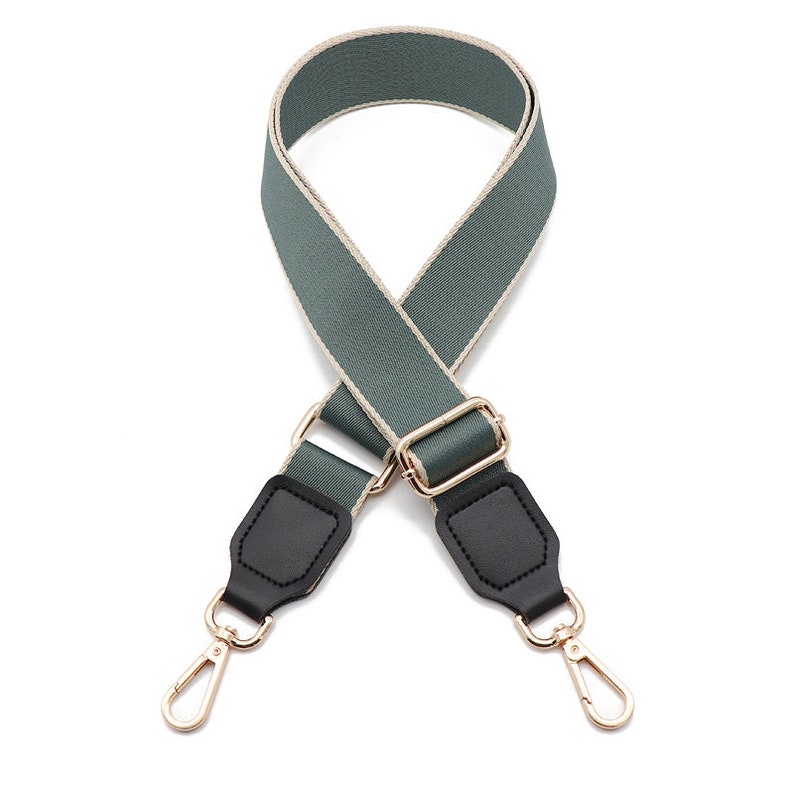 May include: A sage green adjustable bag strap featuring a woven texture with light beige edges. The strap includes black faux leather tabs at both ends, connected to gold-toned metal hardware, including a sliding buckle and two swivel lobster clasps.