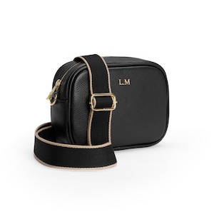 May include: A black pebbled leather crossbody bag with gold-tone hardware and a thick black fabric strap featuring beige trim. The front of the bag is embossed with the gold letters L.M. The bag has a top zipper closure and an adjustable strap buckle.