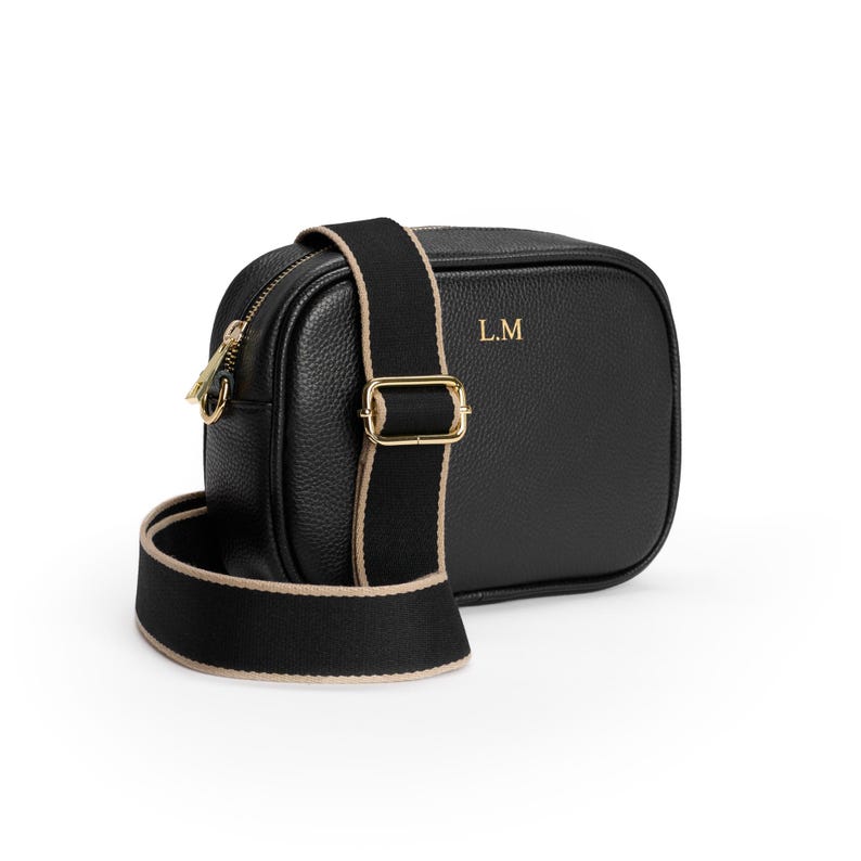 May include: A black pebbled leather crossbody bag with gold-tone hardware and a thick black fabric strap featuring beige trim. The front of the bag is embossed with the gold letters L.M. The bag has a top zipper closure and an adjustable strap buckle.