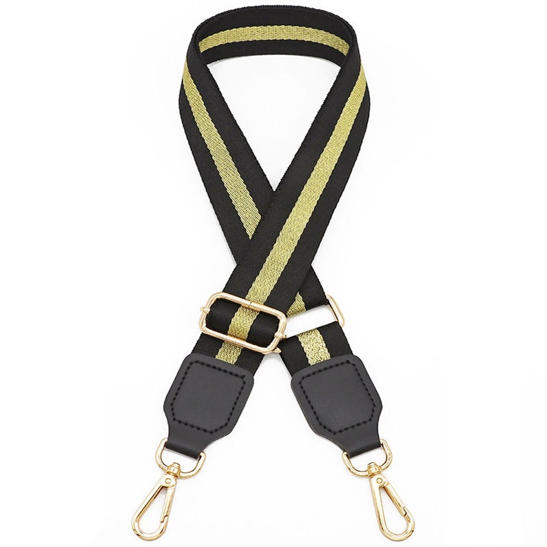 May include: A replacement bag strap featuring a black and gold woven pattern with a central gold stripe. The strap includes a gold metal adjustable slider and two black faux leather end tabs, each finished with a gold metal swivel lobster clasp.